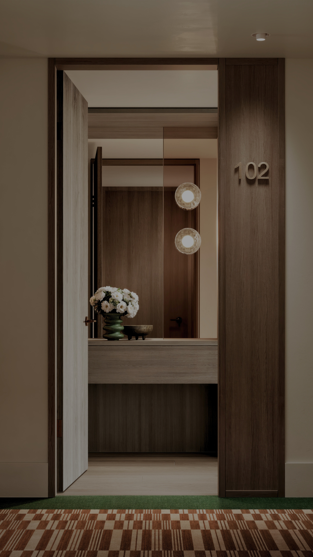 A modern hotel room entrance labelled 102 with a wooden door partially open, revealing a hallway, a console table with white flowers, and two pendant lights—echoing the sophisticated style of luxury flats Melbourne is known for.
