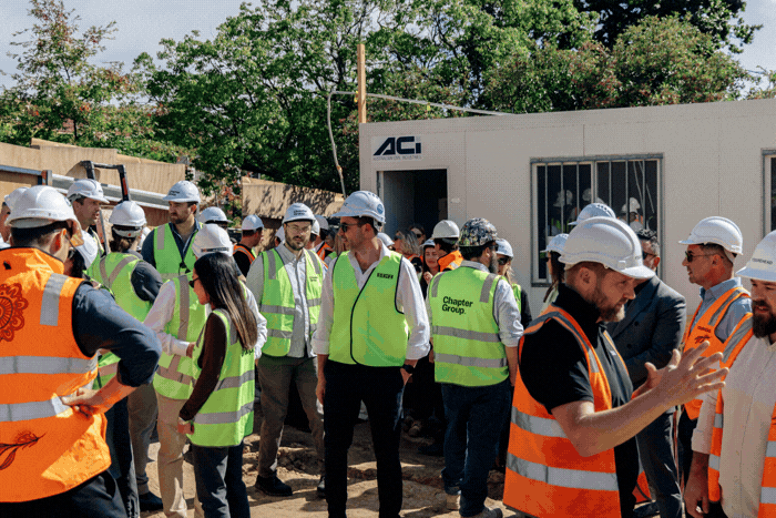 A group of construction workers and professionals in safety vests and helmets gather outside a portable building at the Maleela Rise construction site, engaging in conversation about the forthcoming luxury flats in Melbourne.