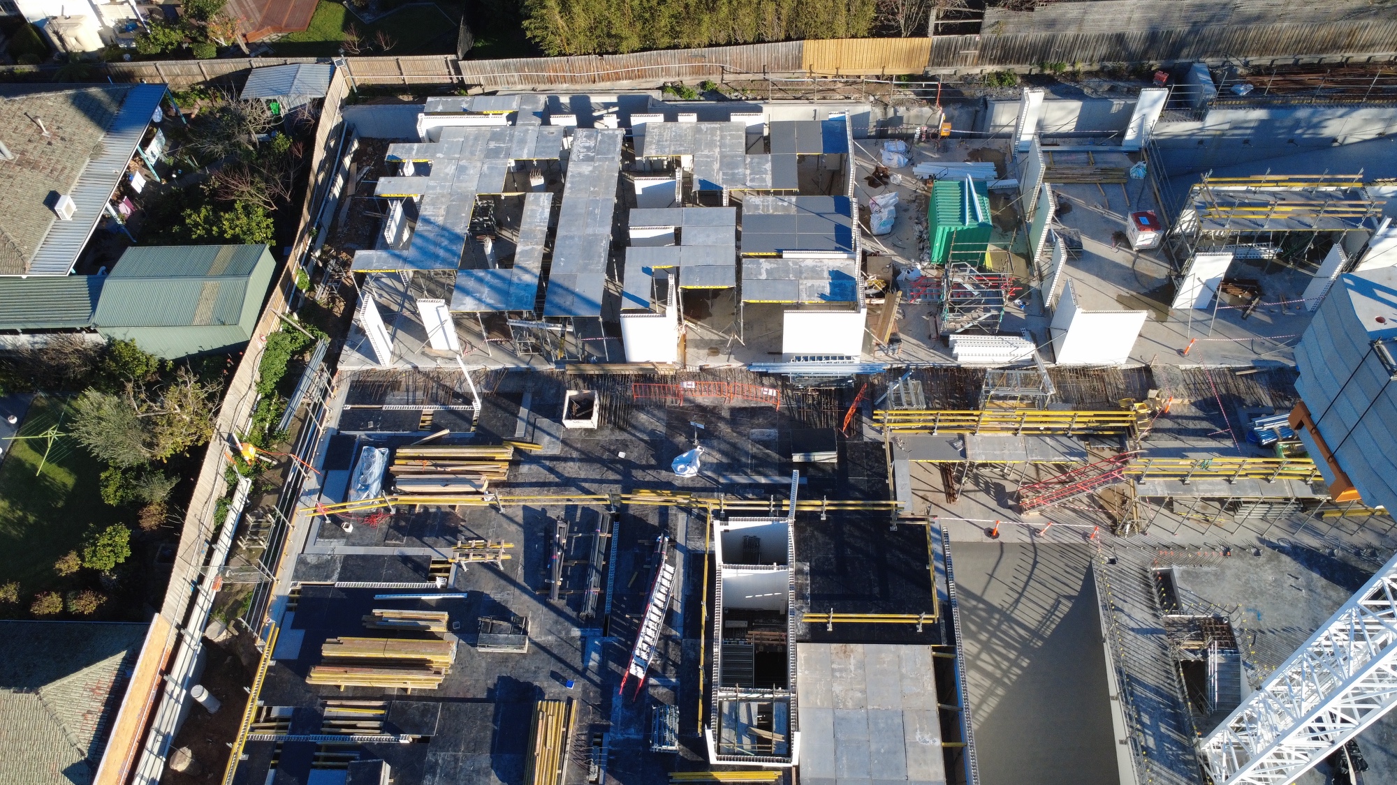 Aerial view of a construction site with partially built concrete structures, equipment, building materials, and scaffolding. Residential houses and trees border the site.