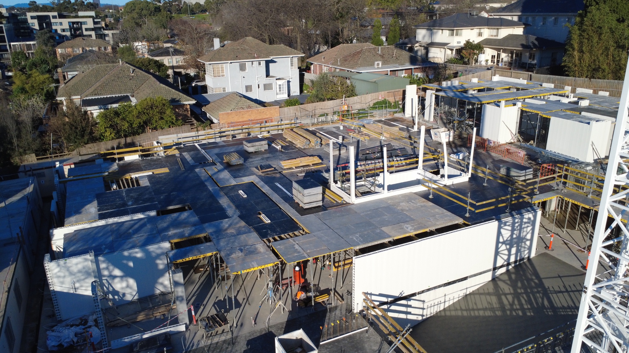 Aerial view of a construction site with concrete floors, steel framework, scattered materials, and partially built walls. Residential houses and trees surround the work area.