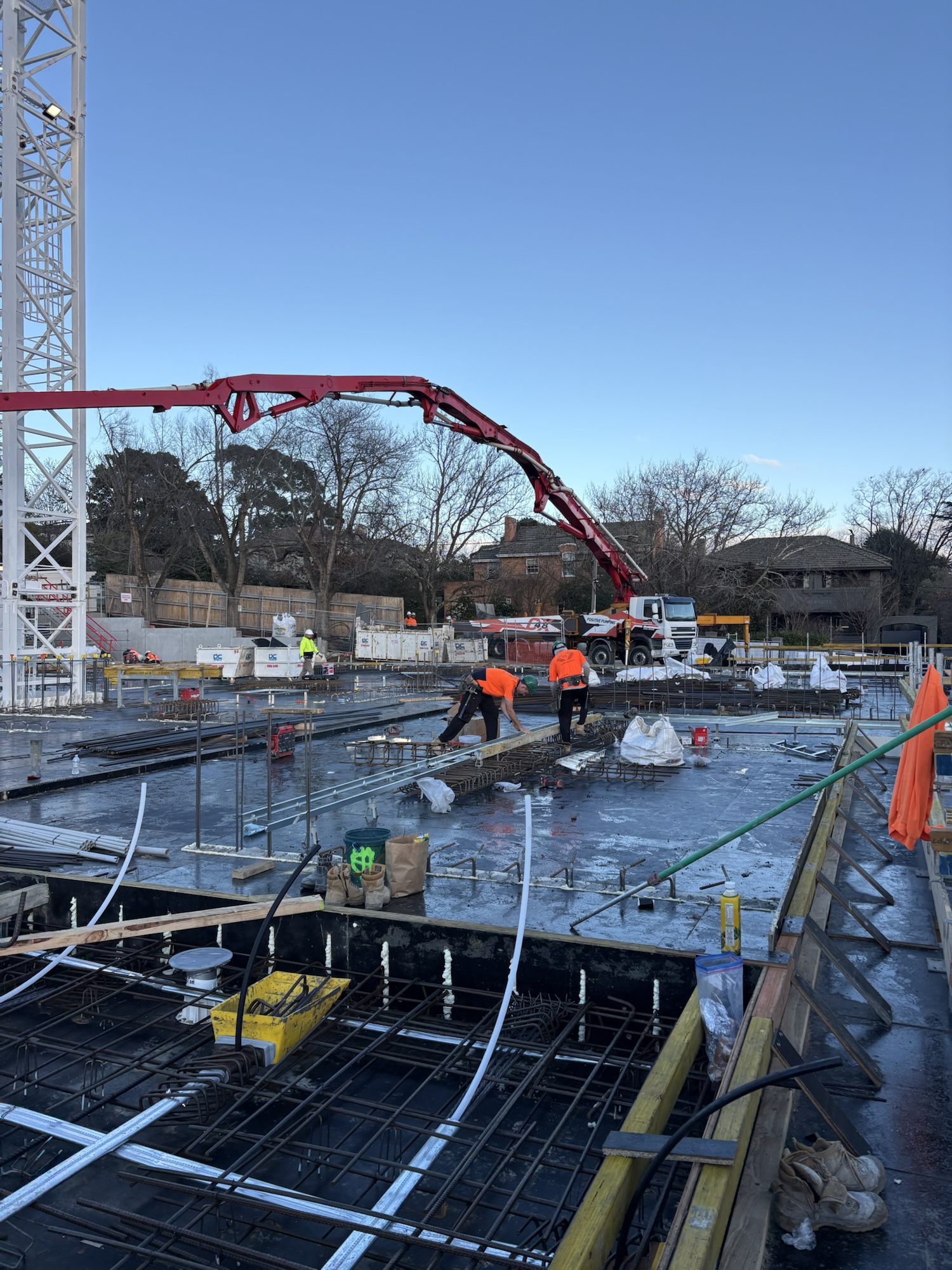 Construction workers in orange jackets pour concrete on a building site, with a red concrete pump and steel reinforcement bars visible. Trees and houses are in the background under a clear blue sky.
