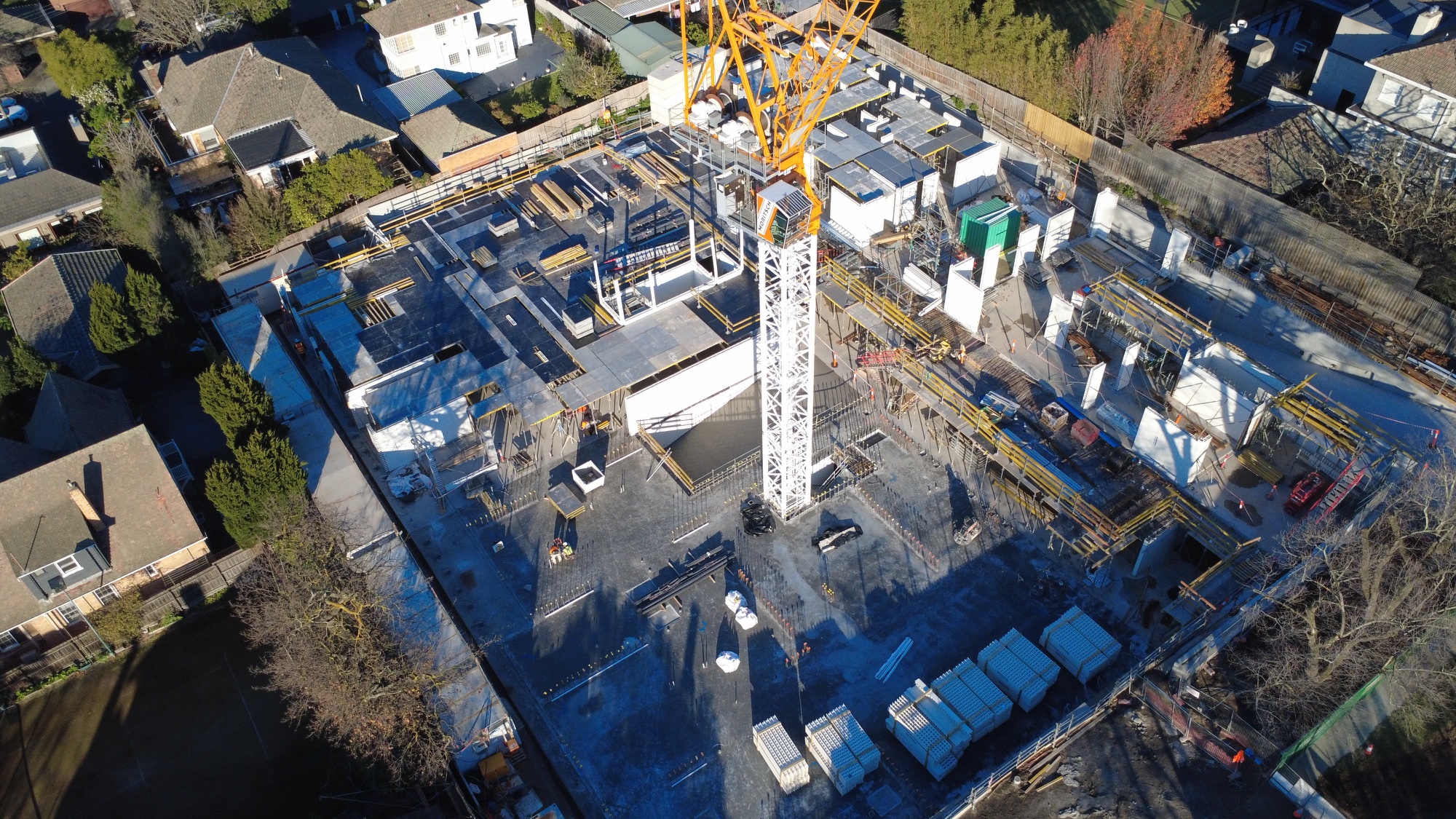 Aerial view of a construction site with a large crane, workers, building materials, and partially built structures, surrounded by residential houses and trees.