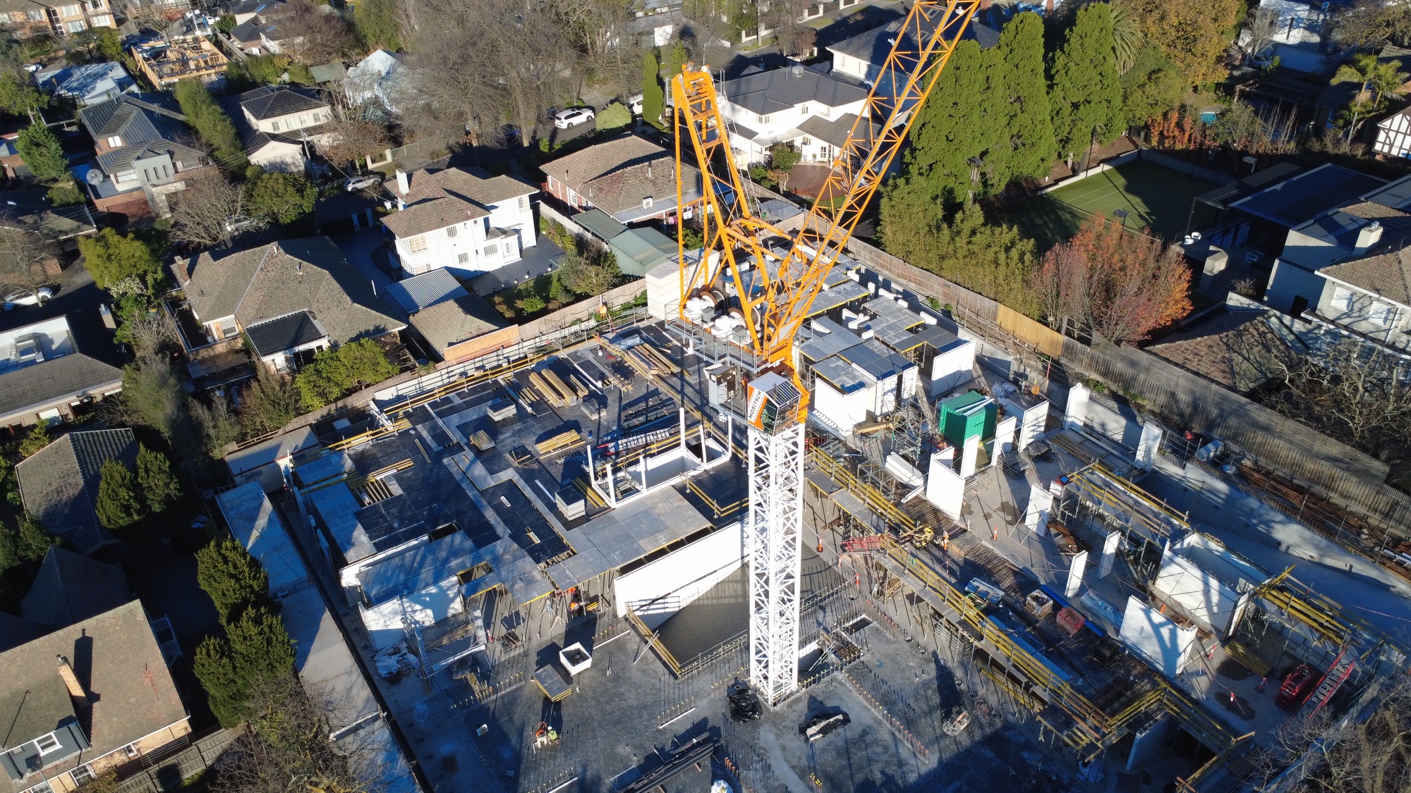 Aerial view of a construction site with a large crane in the centre, surrounded by houses and trees in a residential neighbourhood. Concrete foundations and structures are visible.