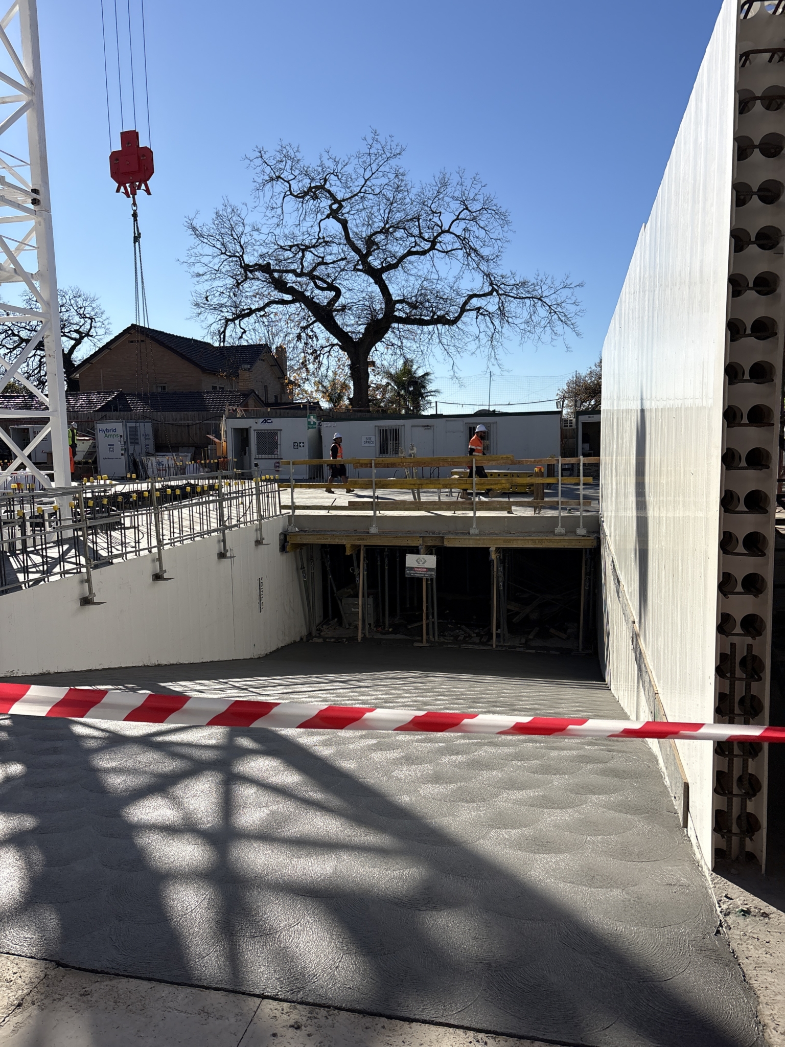 Fresh concrete covers the ground at a construction site, cordoned off with red and white tape. Workers stand on a raised platform, with trees and a crane visible in the background under a clear blue sky.