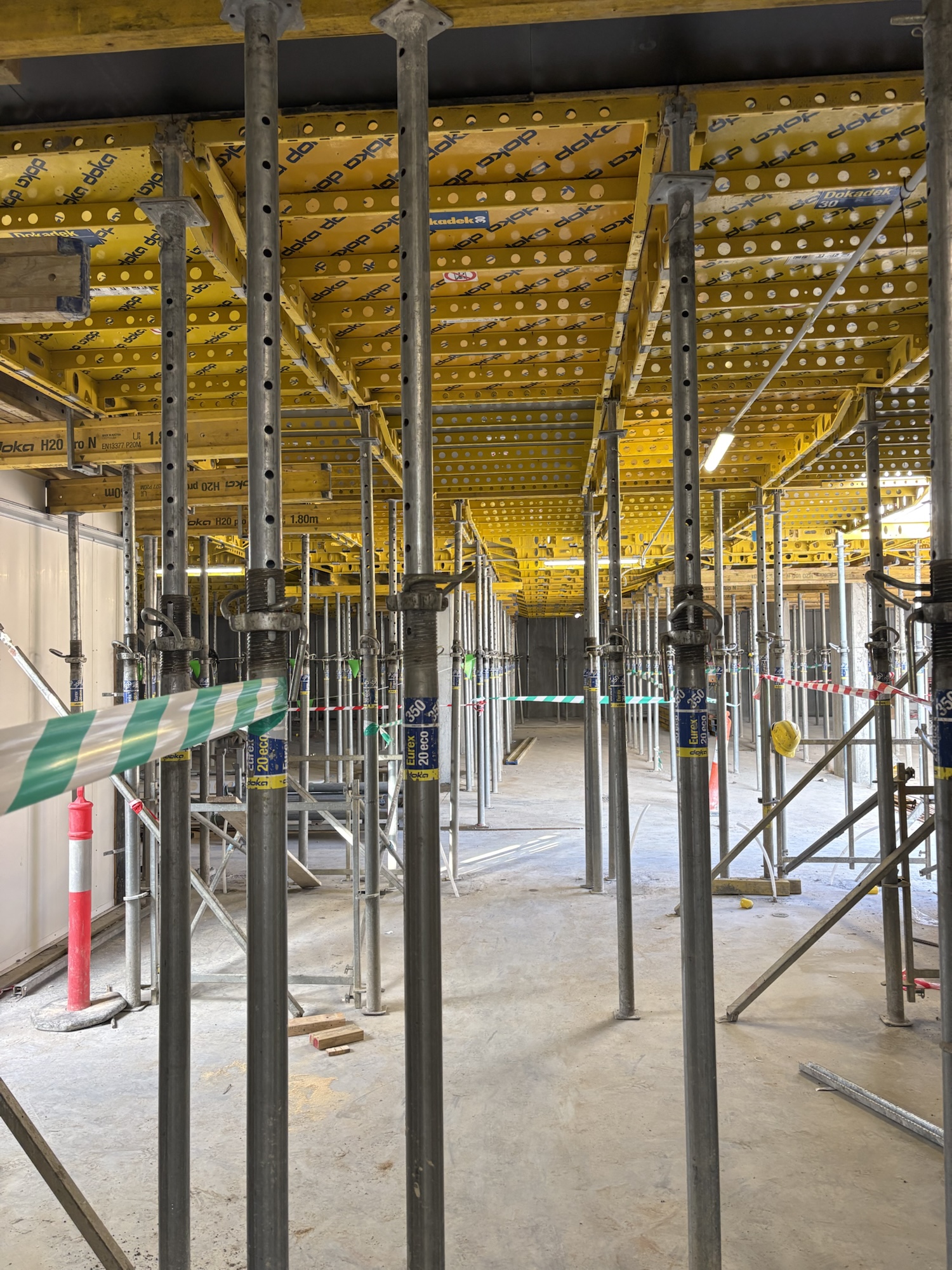 Rows of metal scaffolding poles support a yellow ceiling at a building site. Green and white striped tape blocks part of the area. The space is unfinished, with concrete floors and no walls.