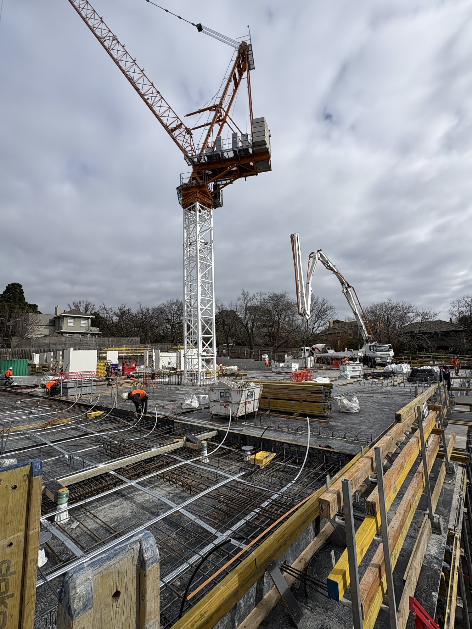 A building site with workers, a tall orange and white crane, concrete pump, and building materials under a cloudy sky. Trees and houses are visible in the background.